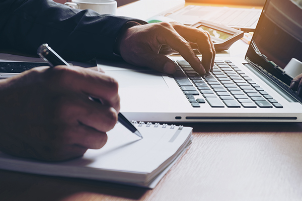 closeup of person writing on paper with a pen, while typing on a laptop keyboard