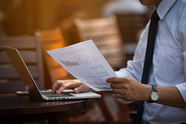 business man holding paper full of data, while typing on a laptop